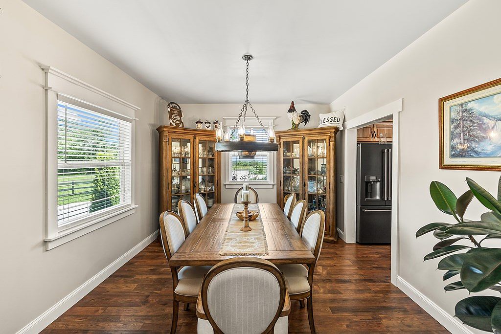Formal dining room with a long farmhouse table seating eight, twin lighted china cabinets, hardwood floors, and tall windows facing the fenced pasture.
